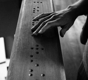 Black & white photograph of a hand reading wood-carved braille code. Author: Christophe MOUSTIER - 2005
