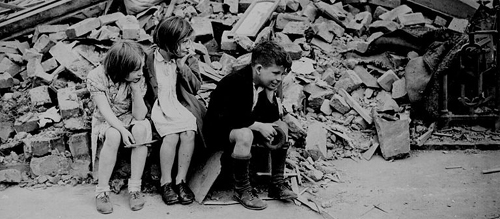 Children in the east end of London, made homeless by the random bombs of the Nazi night raiders, waiting outside the wreckage of what was their home. September 1940 (National Archives) Author US Government